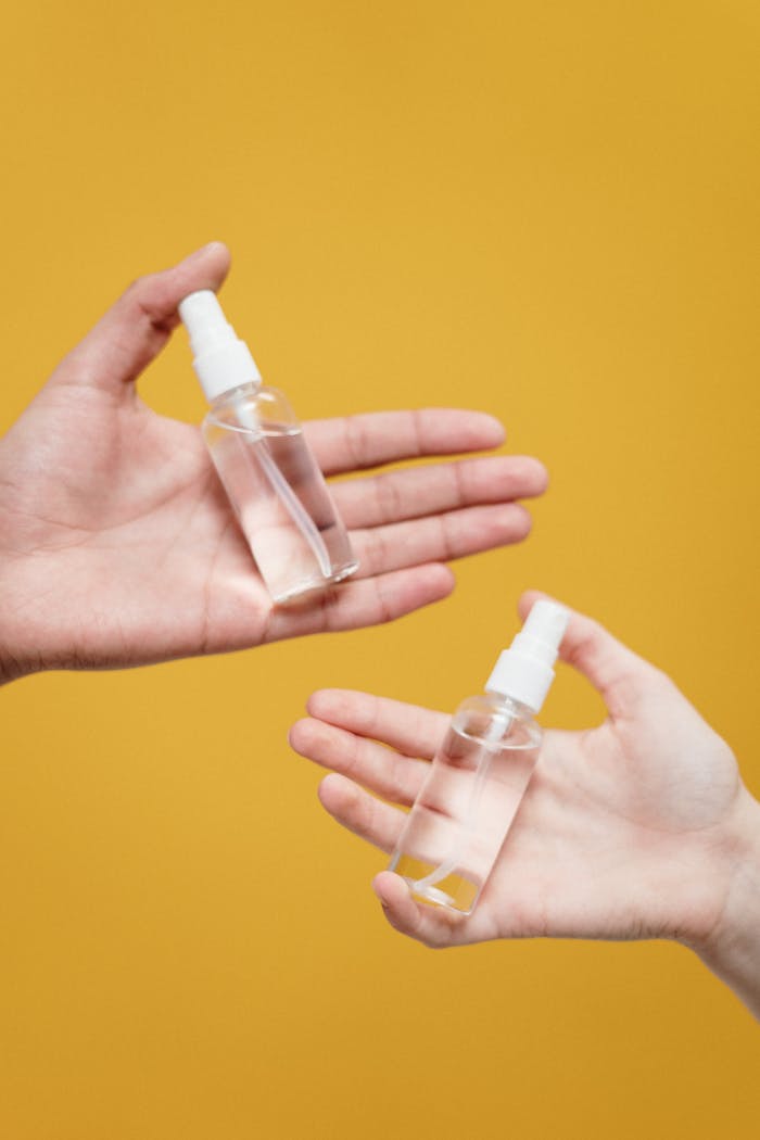 Close-up of two hands holding clear spray bottles on a vibrant yellow background, symbolizing hygiene and safety.